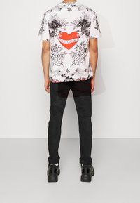 Man som bär en vit T-shirt med svarta blommönster och örn-design, en röd hjärt-grafik som säger "Just Cavalli", svarta byxor och svarta sneakers, står.