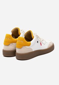 Un paio di sneaker bianche con dettagli in suede giallo sui talloni, accenti beige, suole marroni e un piccolo dettaglio incrociato nero sul lato.