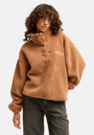 Jeune femme aux cheveux bouclés portant un pull polaire marron et un pantalon noir, debout devant un fond clair uni.