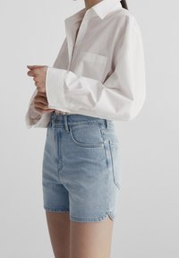 Biała koszula z długim rękawem, zapięta na guziki przy kołnierzyku oraz posiadająca kieszeń na piersi, zestawiona z jasno-niebieskimi szortami dżinsowymi z ozdobnym wykończeniem na brzegu oraz przednimi kieszeniami.