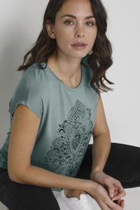 Korte mouwen, teal katoen T-shirt met een ingewikkeld zwart paisleypatroon op de voorkant, gecombineerd met zwarte broek en witte sneakers.