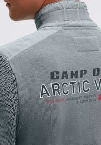 Grauer gestrickter Pullover mit strukturierter Oberfläche, hohem Kragen, gemusterten Schulterakzenten und auffälligem schwarzen und roten Markenaufdruck.