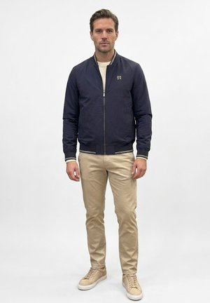 Hombre con chaqueta bomber azul marino sobre pantalones beige y zapatillas beige, de pie frente a un fondo blanco liso.