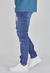 Blue denim jeans with distressed patches, slim fit, mid-rise waist, paired with white sneakers. Fabric texture appears slightly faded.