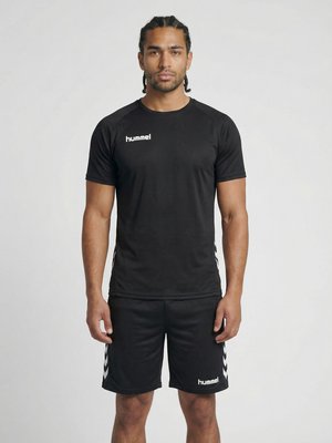 Mannelijk model staand, recht vooruit kijkend, draagt een zwart Hummel sport T-shirt en korte broek tegen een effen grijze achtergrond.