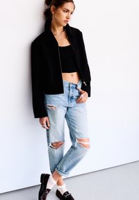 Giacca crop nera con zip su una canotta nera, abbinata a jeans strappati blu chiaro e scarpe nere, con calzini bianchi e anelli d'argento.