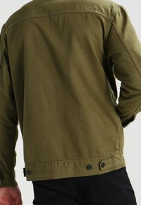 Veste verte olive, en matière de coton doux, avec une fermeture par bouton en bas et des coutures simples le long des coutures. Design classique.