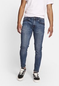 Vaqueros de denim azul ajustados con un ligero desvanecimiento, cinco bolsillos y un cierre de botón. Combinados con zapatillas bajas en blanco y negro.