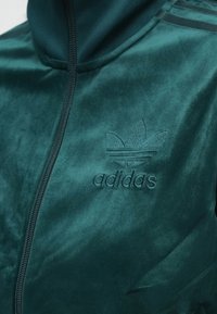 Velvet green zip-up jacket with high collar, featuring embroidered Adidas logo and three striped accents on shoulder. Soft texture.