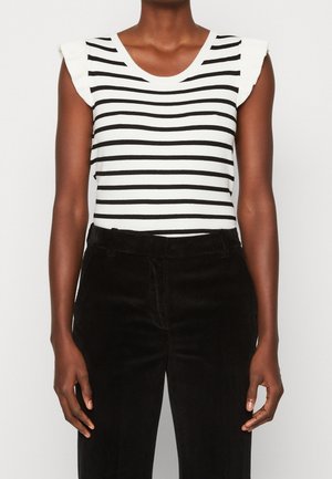 Personne portant un haut blanc sans manches à rayures horizontales noires et un pantalon en velours côtelé noir, debout devant un fond uni.