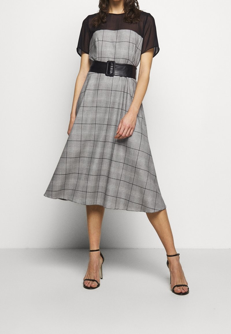 Femme portant une robe à carreaux grise mi-longue avec des manches noires transparentes, une large ceinture noire et des sandales à talons hauts noires.