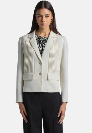 Femme portant une veste blanche cintrée avec des plis verticaux, par-dessus un haut à motifs noir et blanc et un pantalon noir, debout, face à l'avant.