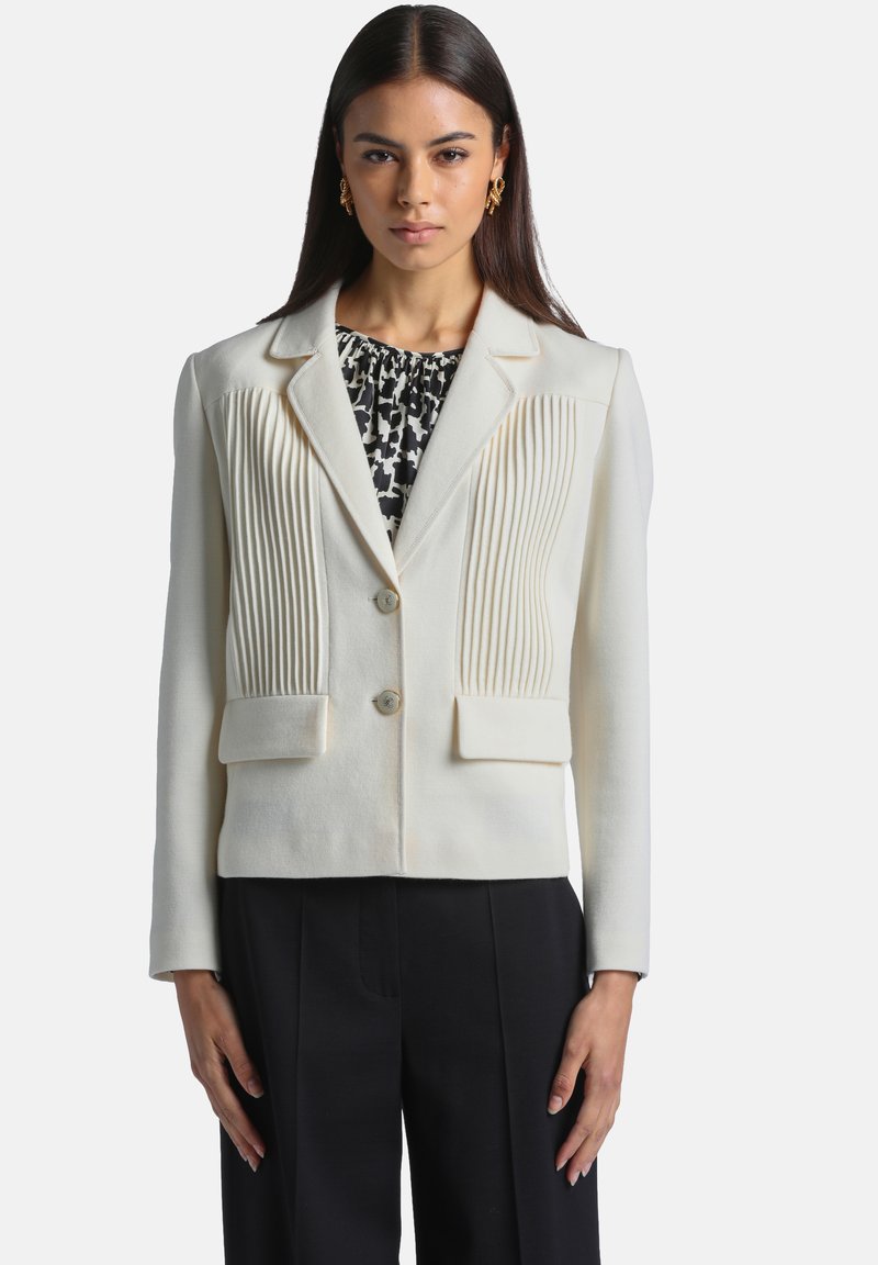 Femme portant une veste blanche cintrée avec des plis verticaux, par-dessus un haut à motifs noir et blanc et un pantalon noir, debout, face à l'avant.