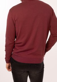 Burgundská pletená mikina s vysokým límcem a žebrovanými manžetami, v kombinaci s černými úzkými džíny. Hladký povrch a těsně přiléhající střih.