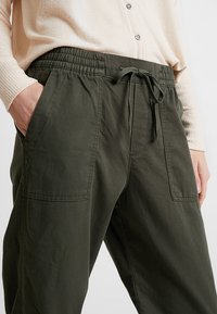 Persoon in een donkergroene broek met voorzakken en een trekkoord in de taille, gecombineerd met een lichtbeige trui.