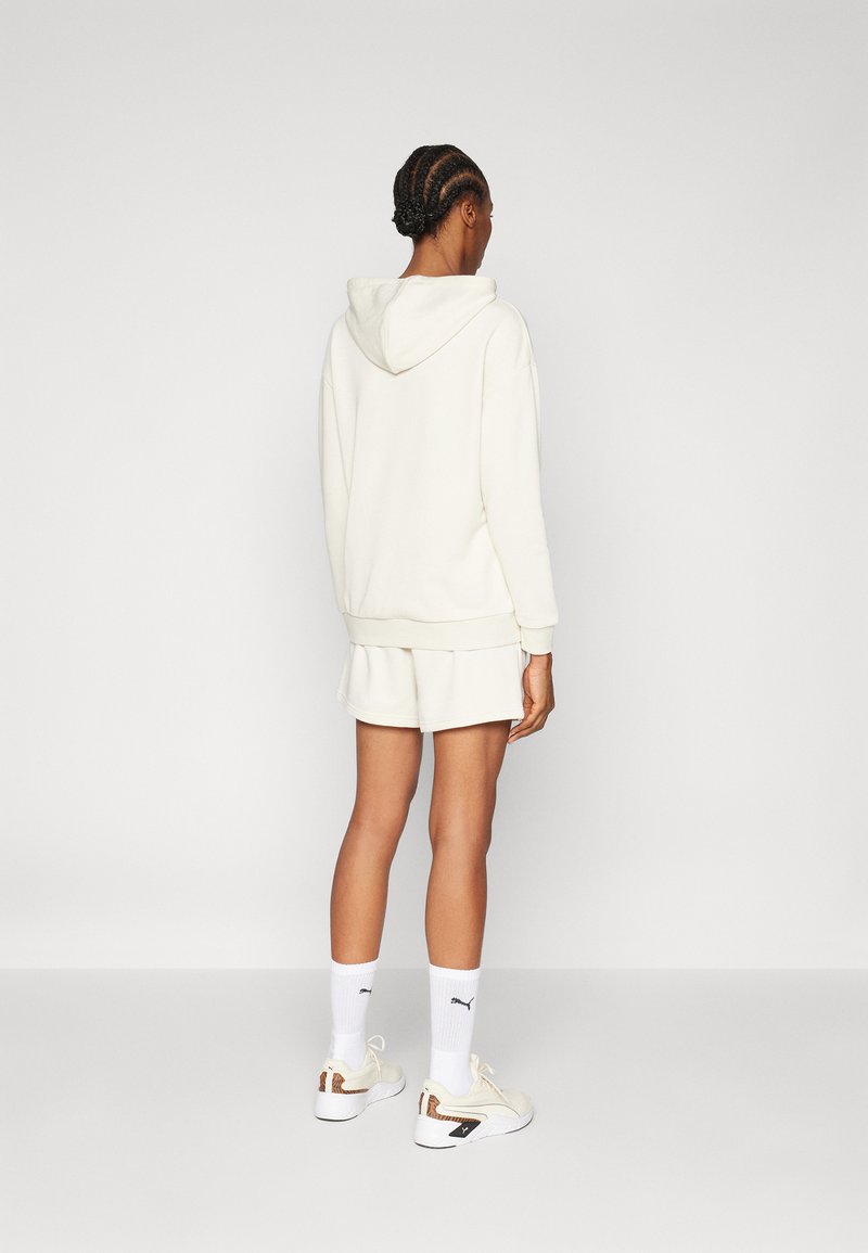 Weiße Kapuzen-Sweatshirt und passende Shorts, aus weichem Material. Kombiniert mit weißen Sneakern und gerippten weißen Socken. Einfaches, minimales Design.