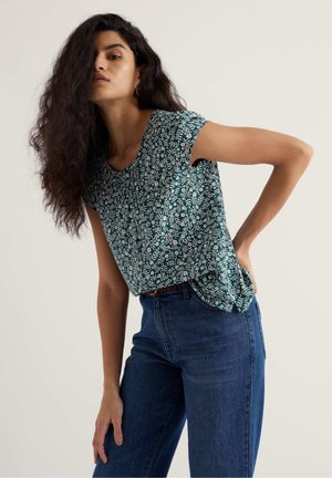 Femeie cu păr lung și creț purtând o bluză cu mâneci scurte, florală, de culoare teal, băgată într-o pereche de blugi albaștri cu talie înaltă, pozând cu o mână pe șold.