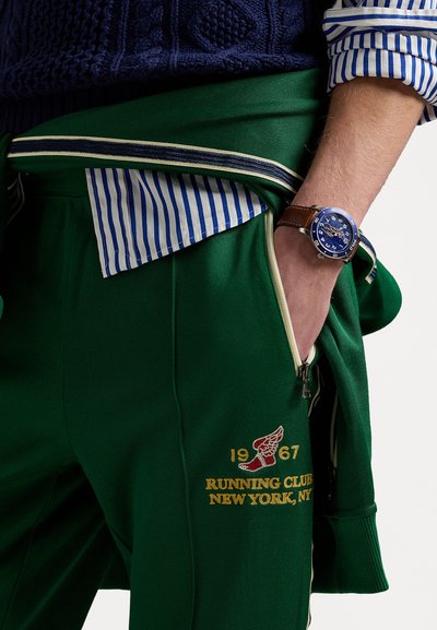 Pantaloni de jogging verzi cu un buzunar cu fermoar, brodați cu "RUNNING CLUB 1967 NEW YORK, NY" în aur, asortați cu un pulover tricotat bleumarin și o cămașă cu dungi.