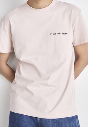 Hellrosa Baumwoll-T-Shirt mit Rundhalsausschnitt und kurzen Ärmeln. Mit schwarzem "Calvin Klein Jeans" Logo auf der linken Brust.