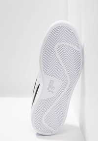 Semelle blanche de basket avec un motif de crampons en zigzag et le logo Puma, positionnée verticalement contre une surface d'angle blanche.