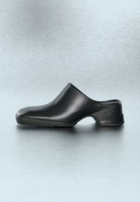 Κομψό μαύρο clog slip-on με λεία επιφάνεια, στρογγυλεμένη μυτερή άκρη και χαμηλό τακούνι τύπου block, εμφανίζεται σε ανοιχτό γκρι φόντο.