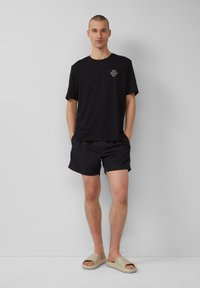 Schwarzes T-Shirt mit kurzen Ärmeln und Logo, kombiniert mit schwarzen Shorts und beigen Schlappen. Glattes Material, lockerer Schnitt und minimalistisches Design.