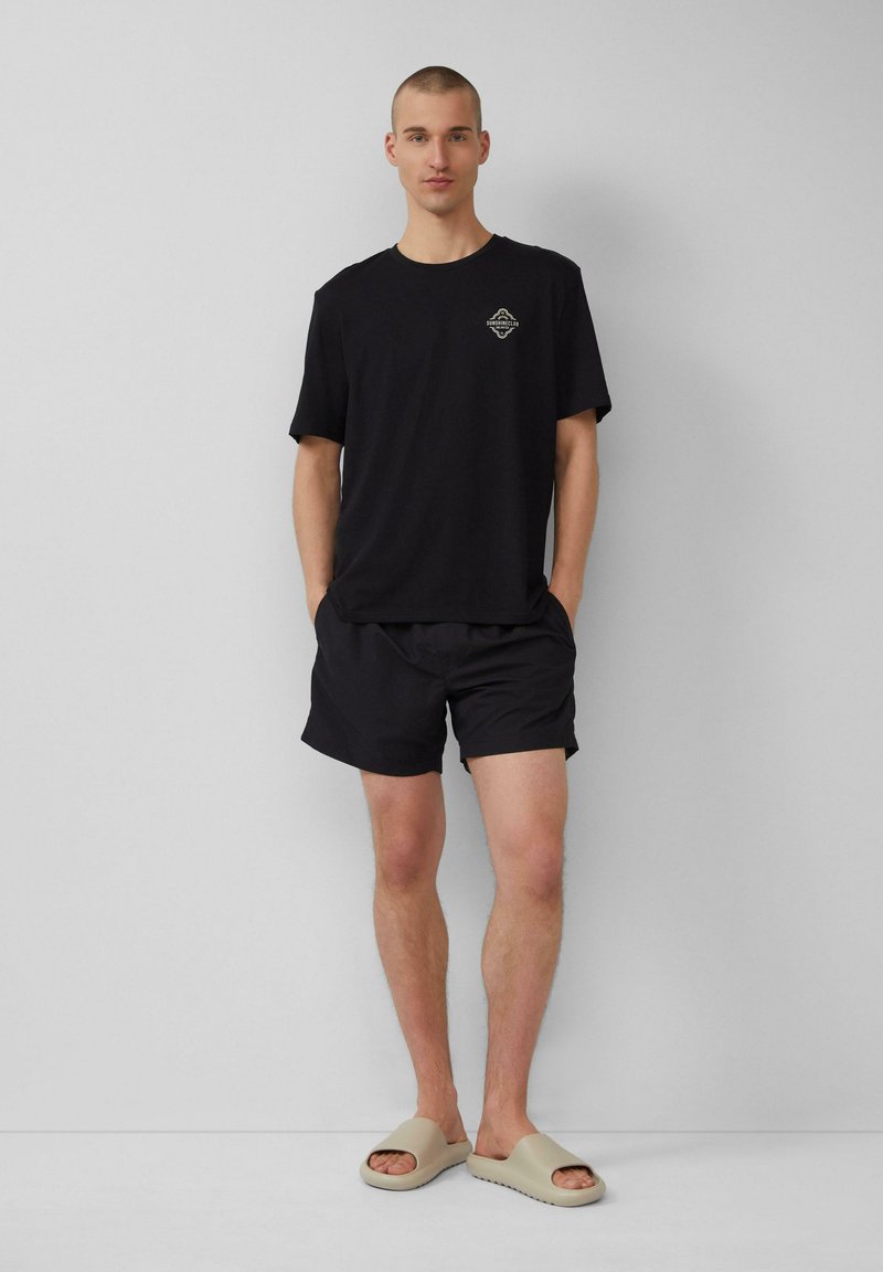 Schwarzes T-Shirt mit kurzen Ärmeln und Logo, kombiniert mit schwarzen Shorts und beigen Schlappen. Glattes Material, lockerer Schnitt und minimalistisches Design.