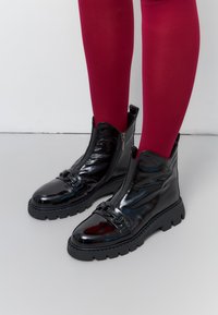 Zwarte gepatineerde enkellaarsjes met een ritssluiting, stevige zwarte zool en decoratieve gespdetails. G gedragen met rode maillots.