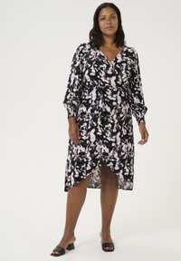 Vestido negro con patrón abstracto rosa y blanco, escote en V, mangas largas abullonadas y dobladillo asimétrico. Usado con sandalias de slid negras.
