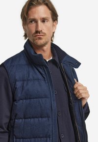 Marineblaue Steppweste mit hohem Kragen, gestepptem Design und Frontreißverschluss. Wird über einem dunklen Hemd mit Knöpfen und sichtbarem Innenfutter getragen.