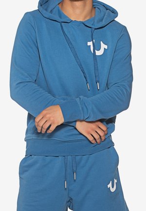 Persoon draagt een blauwe hoodie en bijpassende blauwe joggingbroek met wit logo, armen gekruist ter hoogte van de taille, met zichtbare trekkoorden aan beide kledingstukken.