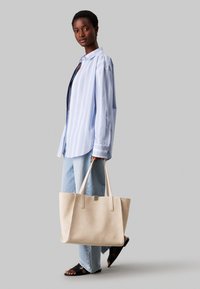 Beige ruskinds tote bag med lange håndtag, rektangulær form og minimal detaljer. Modellen bærer en lyseblå stribet skjorte og denim jeans.