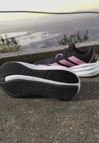 Czarne i różowe buty sportowe na asfalcie, jeden pokazuje wzór bieżnika, z rozmytym panoramą miasta i zielenią w tle.