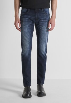 Dark blue denim jeans with a slim fit, featuring a faded finish, five pockets, and a traditional belt loop design, paired with black boots.