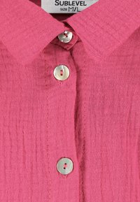 Roze overhemd met knoopsluiting van een geprofileerde stof. Bevat een kraag en drie glanzende zilveren knopen aan de voorkant. Maatlabel zichtbaar.