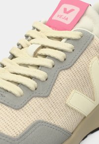 Μπεζ αθλητικό παπούτσι με γκρι σουέντ διακοσμητικά, επάνω μέρος από υφασμάτινη ύλη με υφή, κρέμ κορδόνια και ροζ ετικέτα με το λογότυπο «VEJA» στη γλώσσα του παπουτσιού.
