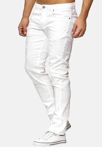 Jean en denim blanc présentant des détails effilochés, une coupe slim, et une fermeture éclair avec bouton, associés à des baskets blanches.