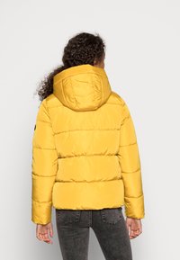 Žlutá vycpaná bunda s kapucí, se štepovanými částmi a předními kapsami. Materiál vypadá hladce a leskle.