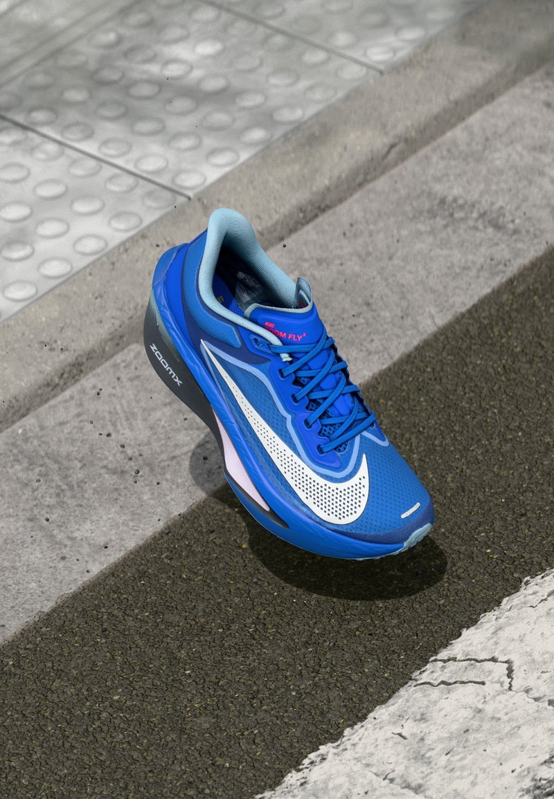 Chaussure de course Nike ZoomX bleue avec un swoosh blanc sur une surface asphaltée près d'un revêtement tactile et d'un trottoir.
