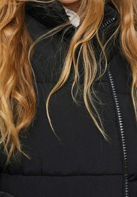 Czarna pikowana kurtka z wysokim kołnierzem, z przodem na zamek błyskawiczny i gładką teksturą, częściowo zasłonięta długimi, falującymi włosami.