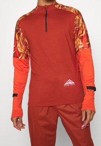 Rotes Sportshirt mit langen Ärmeln, Reißverschlusskragen und abstrakt gemusterten Schultern; verfügt über ein Logo am Saum und Daumenöffnungen in den Ärmeln.