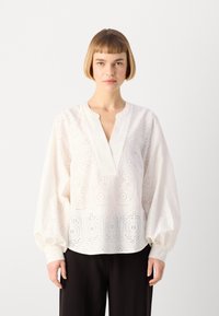 Blusa bianca con ricamo ad occhio di pernice, scollatura a V e maniche lunghe a sbuffo. Vestibilità ampia con motivi intricati. Abbinata a pantaloni neri.