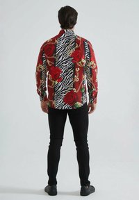 Chemise à boutons multicolore avec un mélange de roses rouges, de chaînes et de rayures zèbres sur le tissu. Manches longues et col pointu. Jean noir porté.