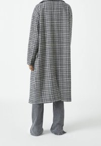 Pitkä harmaa-valkoinen ruudullinen takki, jossa on jämäkkä kaulus ja joka on valmistettu teksturoidusta kankaasta. Yhdistetty harmaisiin levenevöihin farkkuihin, kuvattu takaa.