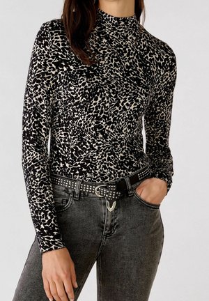 Femme portant un haut à manches longues imprimé animal noir et blanc rentré dans un jean noir délavé avec une ceinture noire cloutée, main dans la poche.