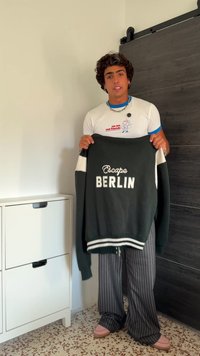 Giovane uomo che tiene in mano una giacca bianca e nera con la scritta "Escape BERLIN", indossando una maglietta bianca con grafica, pantaloni a righe e scarpe rosa all'interno.