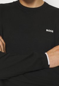 Chemise noire à manches longues avec une texture côtelée. Présente un logo "BOSS" blanc sur la poitrine et un bord blanc contrastant aux poignets des manches.