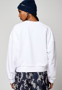 Felpa bianca con collo a giro, spalle cadenti, polsini e orlo ribattuti. Indossata sopra pantaloni scuri a fantasia, completata da un berretto blu.