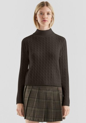 Jeune femme portant un pull à col roulé en maille torsadée marron foncé et une jupe plissée à carreaux marron, debout devant un fond uni.