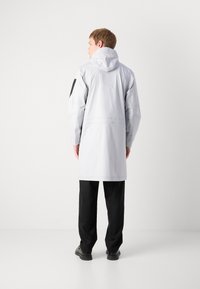 Jachetă impermeabilă de culoare gri deschis, cu glugă, mâneci lungi și un accent negru pe brațul stâng. Design lejer, până la genunchi, cu o textură netedă.